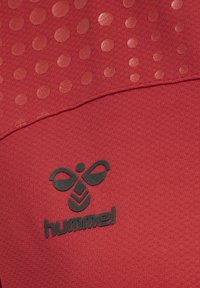 Roter Sportstoff mit strukturierten erhabenen Punkten und einem schwarzen hummel-Logo, das nahe einer geschwungenen Naht gedruckt ist.