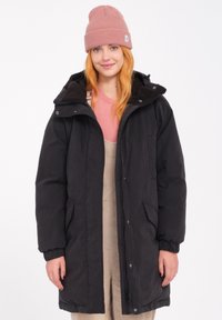 Zwarte pufferjack met een rits aan de voorkant, zijzakken en een capuchon. Het model draagt het over een beige outfit en een roze muts. Gladde textuur.