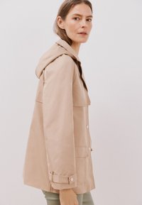 Beige regnjacka med huva, med stora kragar, dolda knappar och sidofickor, designad med en något figurnära passform och slät textur.