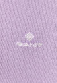 Fialová bavlněná látka s texturovanou vazbou; bílé vyšívané logo s nápisem "GANT" a diamantovým tvarem nad ním.