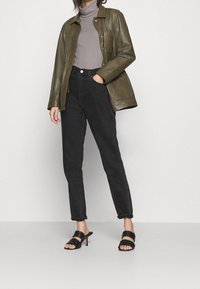 Olivegrüne Lederjacke mit Taschen, grauer Rollkragenpulli, schwarze hochgeschnittene Jeans und schwarze Sandalen mit Absatz und zwei Riemen.