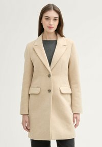 Beige ullblandad kappa med skräddarsydd design, knapphålskrage, två framfickor och en enkel knappstängning. Slät textur.
