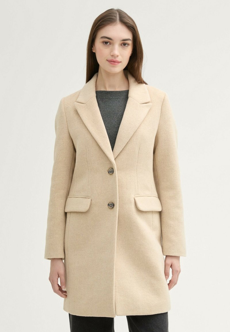 Cappotto in misto lana beige con un design su misura, rever a punta, due tasche frontali e chiusura con un solo bottone. Texture liscia.