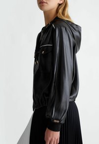 Giacca nera in satin con cappuccio, dotata di due tasche frontali, dettagli in oro e polsini elasticizzati. Texture liscia, design lineare.