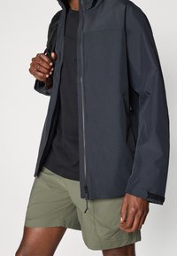Veste imperméable navy avec capuche, fermeture éclair à l'avant et poignets ajustables, assortie à un t-shirt noir et un short vert olive.