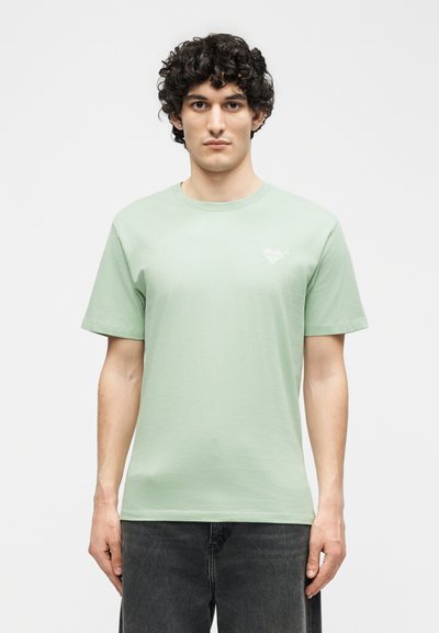 Νεαρός άνδρας με σγουρά σκούρα μαλλιά που φοράει ανοιχτοπράσινο κοντομάνικο μπλουζάκι με μικρό σχέδιο καρδιάς στο στήθος.