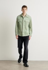 Cămașă verde cu dungi, cu nasturi, prevăzută cu două buzunare pe piept, asortată cu pantaloni negri drepți și încălțăminte neagră, pe un fundal neutru.