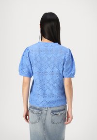 Haut bleu en œillets avec des manches courtes bouffantes, présentant un motif en losanges et des bords festonnés. Associé à une jupe en denim, affichant un look décontracté.
