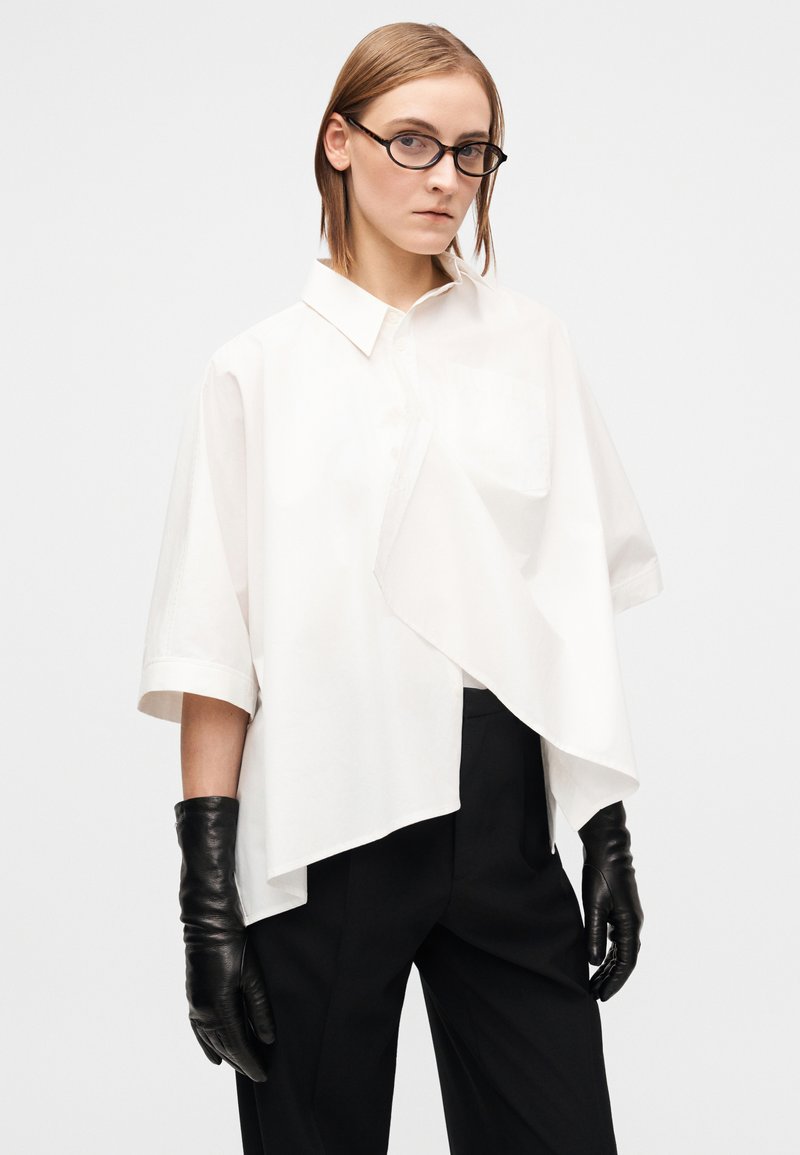 Chemise blanche oversize avec un rabat diagonal à l'avant, manches courtes, une poche poitrine, associée à des gants en cuir noir et un pantalon noir ajusté.