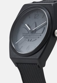 Montre analogique noire avec un fini mat, dotée d'un cadran rond, d'un bracelet noir texturé et d'un design de logo sur le cadran. Aiguilles et index argentés.