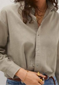 Camicia button-up verde oliva chiaro realizzata in tessuto morbido, con colletto classico e vestibilità rilassata. Dettagli notevoli includono piccoli bottoni e maniche arrotolate.