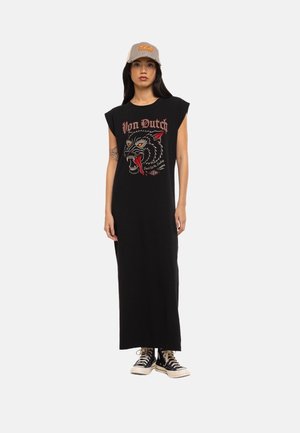 Femme portant une robe maxi noire sans manches avec un motif de loup rouge et blanc et le texte « Von Dutch », casquette beige, baskets montantes noires.