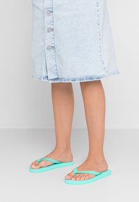 Turquoise flip-flops with a textured sole and thin straps, paired with a light blue denim skirt featuring button details and a frayed hem.