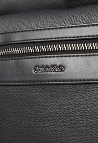Portefeuille en cuir noir présentant une surface texturée, une fermeture éclair argentée et le logo "Calvin Klein" en relief. Un subtil détail de couture ajoute de la structure.