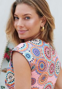 Robe colorée avec un motif floral en orange, rose, bleu et violet. Tissu texturé avec des manches courtes, accentuée par des boucles d'oreilles en or.