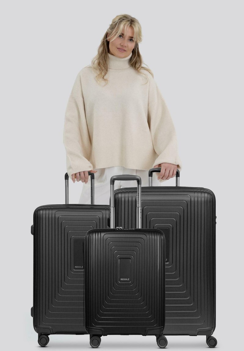 Una donna con capelli biondi che indossa un maglione a collo alto beige tiene in mano le maniglie di tre valigie rigide nere di diverse dimensioni.