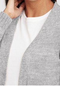 Light gray knitted cardigan with a V-neck, featuring ribbed texture. Worn over a white shirt, showcasing soft fabric and open front design.