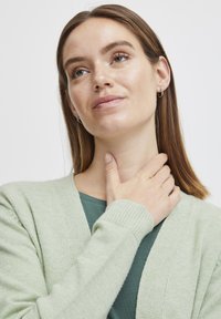 Světlezelený cardigan vyrobený z měkkého materiálu, s volným střihem, žebrovanými manžetami a výstřihem do V, nošený přes tmavozelený top.