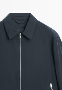 Mørkeblå jakke med spids krave, central sølvlynlås og lynlåslomme på den nederste højre side.