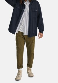 Man som bär en mörkblå knäppt overshirt, randig ljusblå och vit skjorta, olivgröna cargo-byxor, vita strumpor och beige sneakers, står.