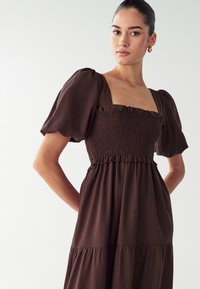 Hnedé šaty s balónovými rukávmi a štvorcovým výstrihom, s vyšívaným telom a viacvrstvovou sukňou. Vyrobené z ľahkej tkaniny s jemnou textúrou.