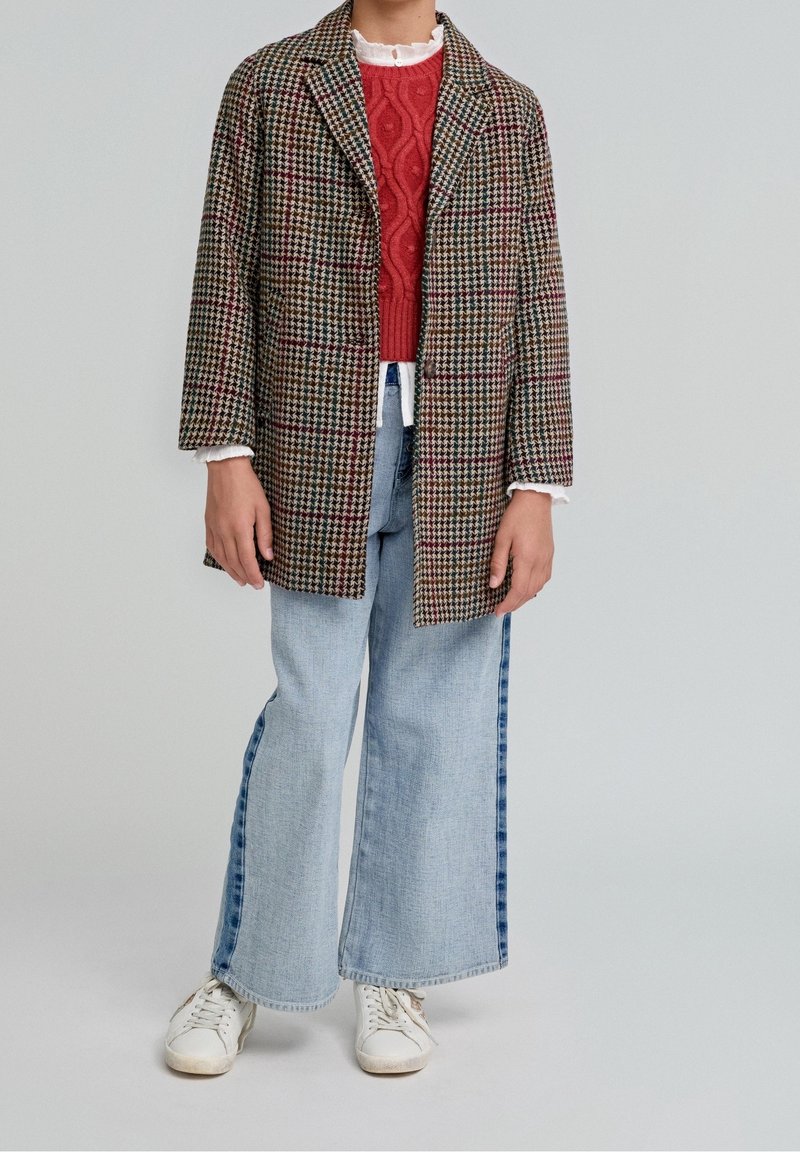 Manteau à carreaux en mélange de laine marron et crème, superposé à un pull rouge en maille torsadée. Associé à un jean large bleu clair et des baskets blanches.