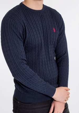 Homme portant un pull en tricot torsadé bleu marine avec un logo rouge brodé sur la poitrine, associé à un pantalon noir, les bras croisés.