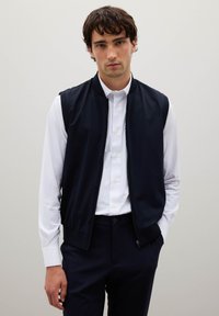 Marineblaues ärmelloses Weste aus leichtem Stoff mit glatter Textur, Frontreißverschluss, geripptem Kragen, kombiniert mit einem weißen Hemd und dunkler Hose.