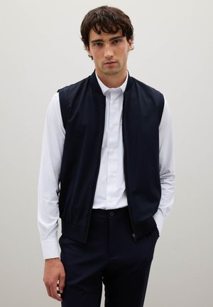 Marineblauw mouwloos vest van lichtgewicht stof met een gladde textuur, ritssluiting aan de voorkant, geribde kraag, gecombineerd met een wit overhemd met knopen en donkere broek.