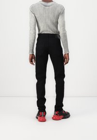 Personne portant un pull gris à manches longues côtelé, un pantalon noir et des baskets noires et rouges, debout sur un fond blanc uni.