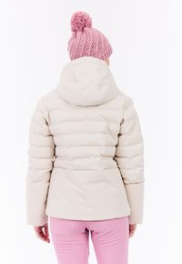 Pufferjack aan in crèmekleur met een getailleerde taille en capuchon, met een gequilt textuur. Combinatie met lichtroze strakke broek en een roze gebreide muts.