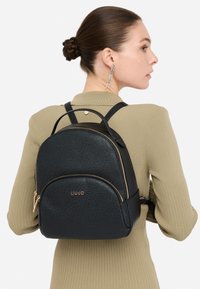 Melns ādas mugursoma ar teksturētu virsmu, zelta krāsas metāla detaļām, apaļu formu un priekšējā daļā rāvējslēdzēja kabatu. Priekšējā daļā izspūdināts logotips.