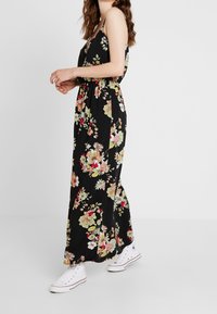 Mönstrad maxi-klänning i svart med livfulla flerfärgade blommor. Lättviktsmaterial, ärmlös design, bärs med vita sneakers.