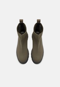 Cizme elastice până la gleznă din material verde măsliniu, cu textură canelată, tălpi din cauciuc negru și taburi pentru prindere la călcâi pentru a fi ușor de îmbrăcat și dezbrăcat.