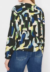 Femme portant un chemisier à manches longues avec des motifs abstraits bleus, verts, jaunes et noirs, associé à un jean denim bleu.