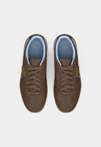 Pruunist sametist tossud, millel on beežid kummist tallad, kerge sinise sisetallaga, ümmargused paelad ning külgedel kollane ja must logo.