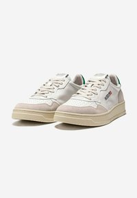 Sneakers basse bianche e beige con punta traforata, chiusura con lacci, linguette verde sul tallone e una piccola toppa rettangolare con il logo sul lato.