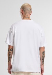 Hvit bomulls T-skjorte med korte ermer og løs passform. Har rund hals og synlige tatoveringer på armer og nakke.