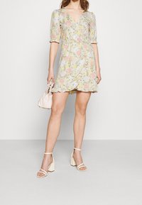 Wickelkleid in leichtem Blumenmuster, mit pastellfarbenen Blüten auf hellem Hintergrund, kurzen Puffärmeln und einem gewellten Saum. Kombiniert mit beigefarbenen Blockabsätzen.