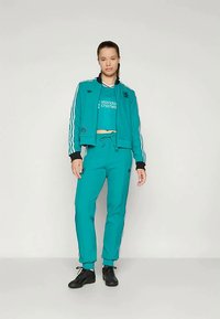 Τιρκουάζ φόρμα με λευκές ρίγες στα μανίκια και στα παντελόνια. Η ζακέτα διαθέτει λαιμόκοψη με πλέξη, ενώ το παντελόνι έχει ρυθμιζόμενη μέση.