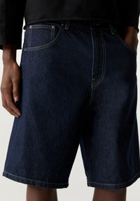 Short en jean bleu foncé à coupe droite, avec surpiqûres jaunes contrastantes, cinq poches et un ourlet roulé aux jambes.