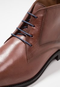 Chaussure habillée en cuir marron avec des lacets bleu marine, couture détaillée et une semelle noire sur fond blanc.