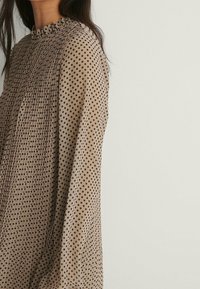 Blusa marrone con scollo arricciato, caratterizzata da pieghe e pois neri. Maniche lunghe con una vestibilità ampia, tessuto texturizzato.