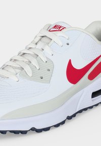Άσπρο παπούτσι Nike με κόκκινο λογότυπο swoosh, λευκά κορδόνια και γκρι λεπτομέρεια στη σόλα, εμφανίζεται από πλάγια γωνία σε απλό φόντο.