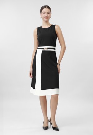 Žena stojaca v čiernych šatách bez rukávov s bielym vertikálnym pruhom a opaskom, doplnených čiernymi lodičkami a zlatými náušnicami.