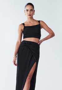 Crni teksturirani crop top s tankim trakama i suknjom visokog struka s detaljem čvora i prorezom do butina, koji prikazuje lepršavu tkaninu.
