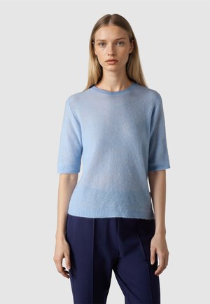 Junge Frau mit glattem blondem Haar, die einen hellblauen Kurzarmpullover und dunkelblaue Hosen trägt, vor einem schlichten Hintergrund stehend.