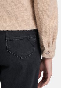 Beżowy sweter z teksturowanym rękawem i guzikowym mankietem obok czarnych dżinsów z kieszenią, ręka luźno opadająca wzdłuż boku na białym tle.