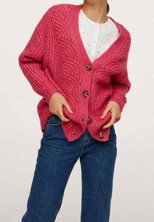 Femme portant un cardigan rose texturé boutonné par-dessus un chemisier blanc avec des détails en broderie et un jean denim bleu.
