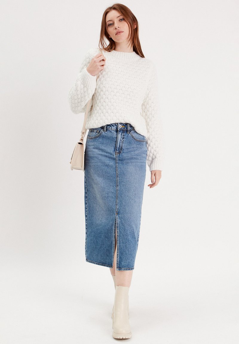 Pull en tricot blanc avec un motif texturé, associé à une jupe midi en denim bleu avec une fente à l'avant. Des bottines beige à la cheville complètent le look.